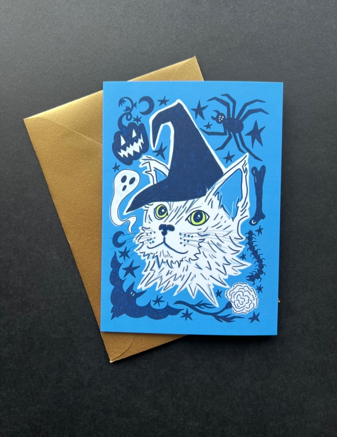 Blue greeting card with a cat wearing a witch hat on a gold envelope against a black background