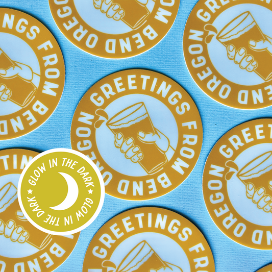 Circular yellow glow in the dark sticker that has an illustrated hand holding a beer with a band of words surrounding the illustration saying “Greetings from Bend Oregon”