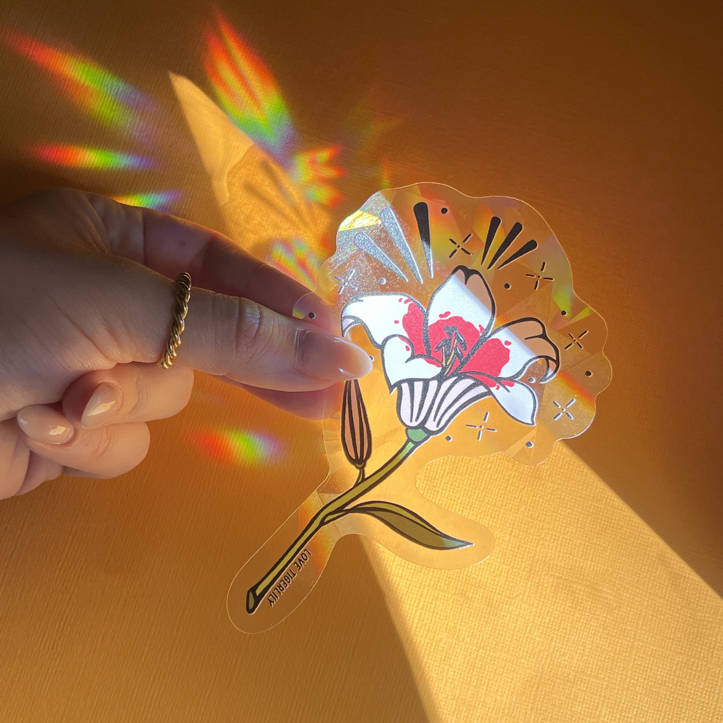 Hand holding the pink and white lily suncatcher window sticker over a yellow background so that it casts rainbows.