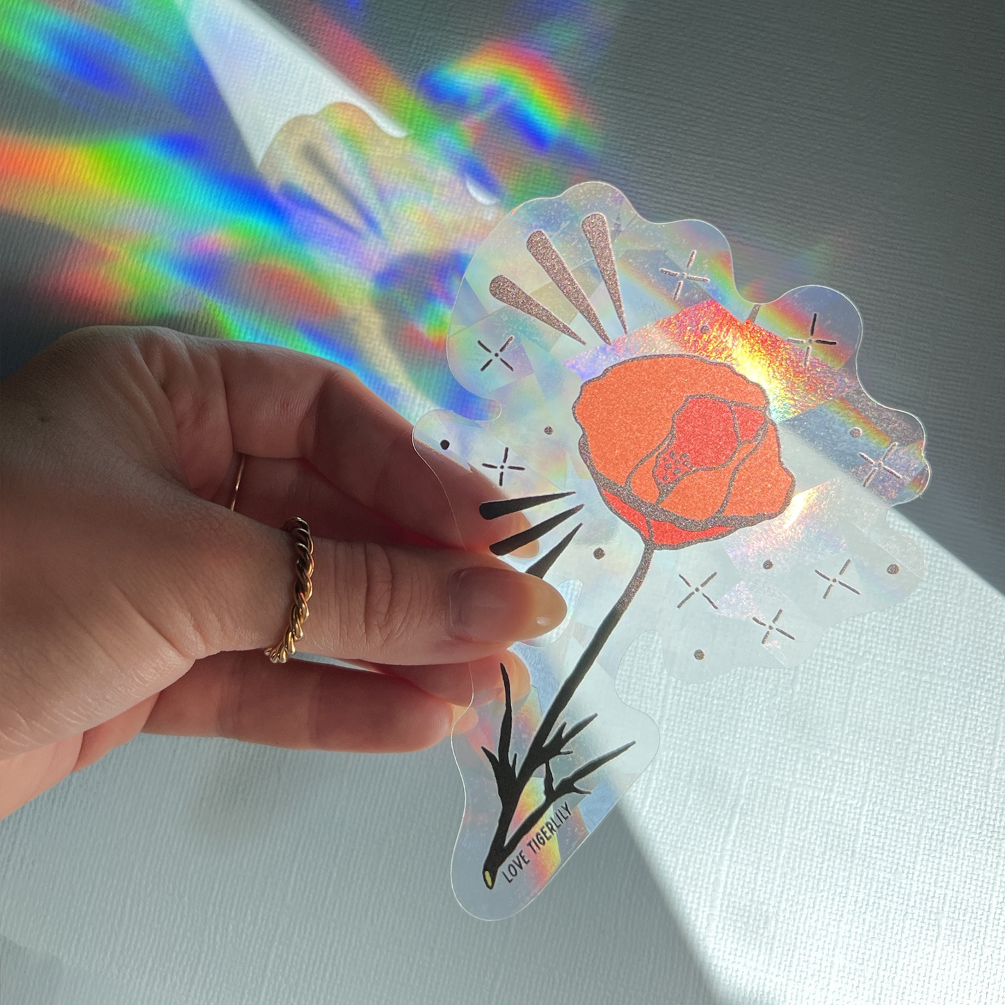 Hand holding the orange California Poppy suncatcher window sticker over a blue background so that it casts rainbows.