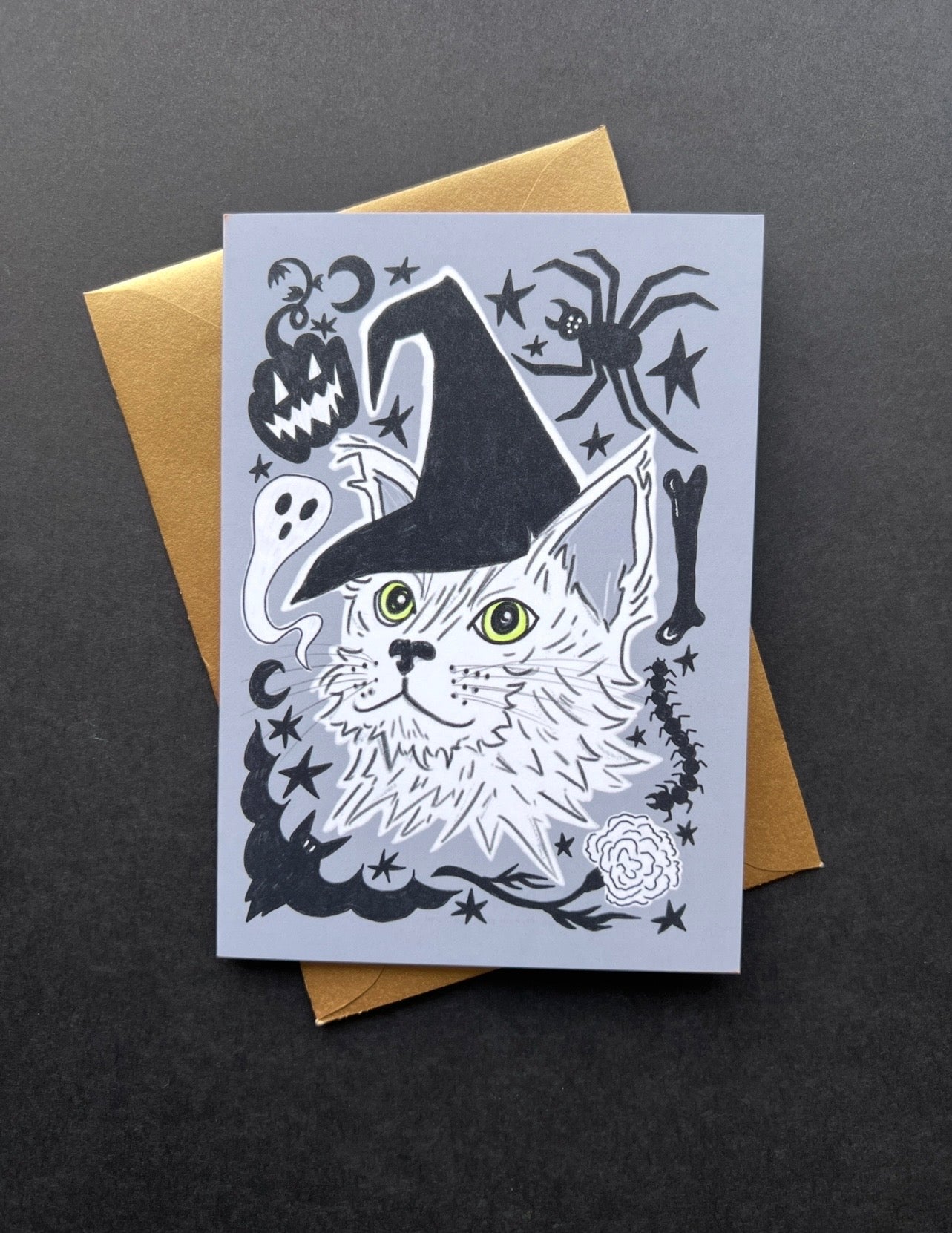 Halloween-themed greeting card with a cat wearing a witch hat on a gray background, placed on a dark surface.