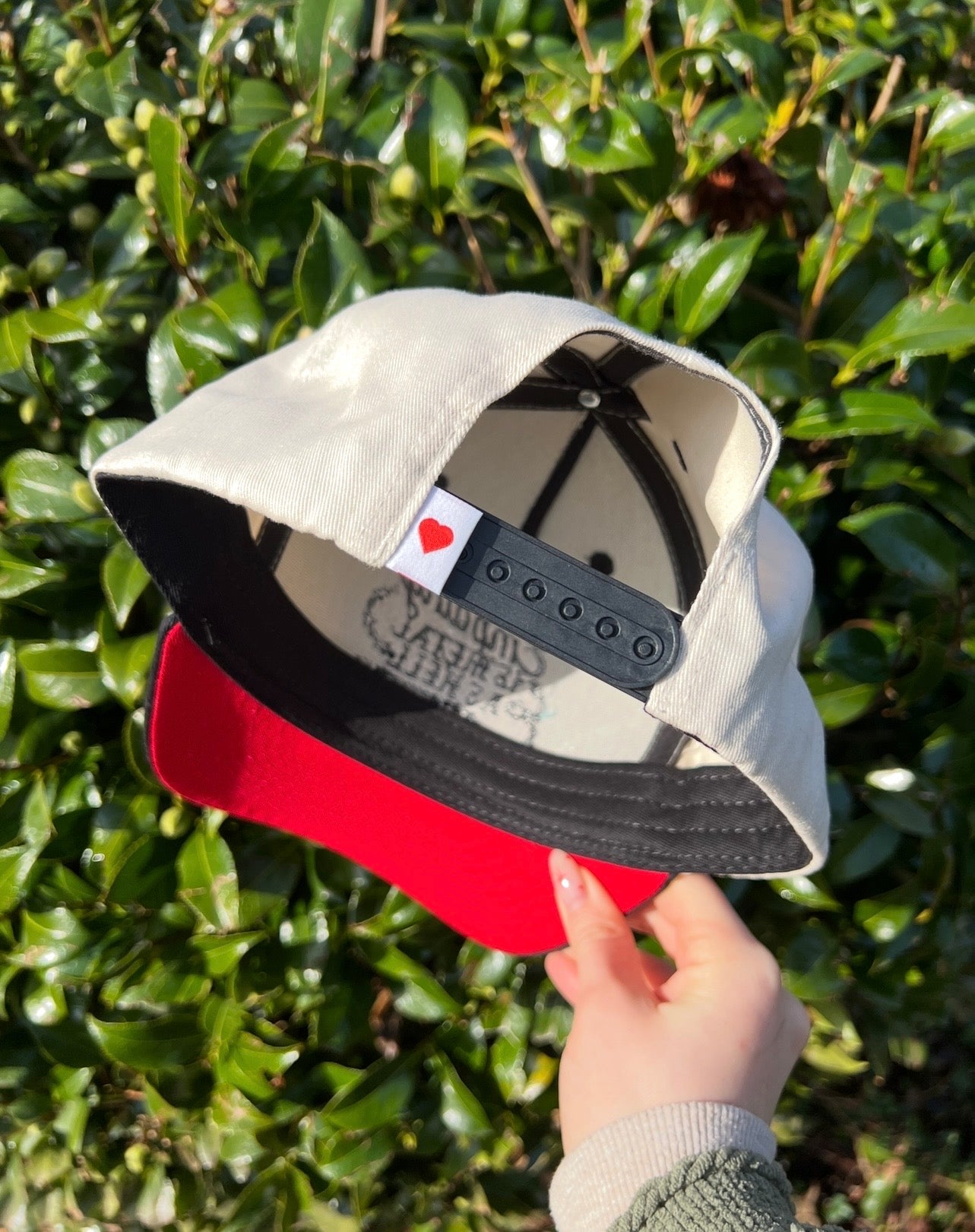 Hand holding a cap with a red brim and an adjustable back with a red heart against a green leafy background