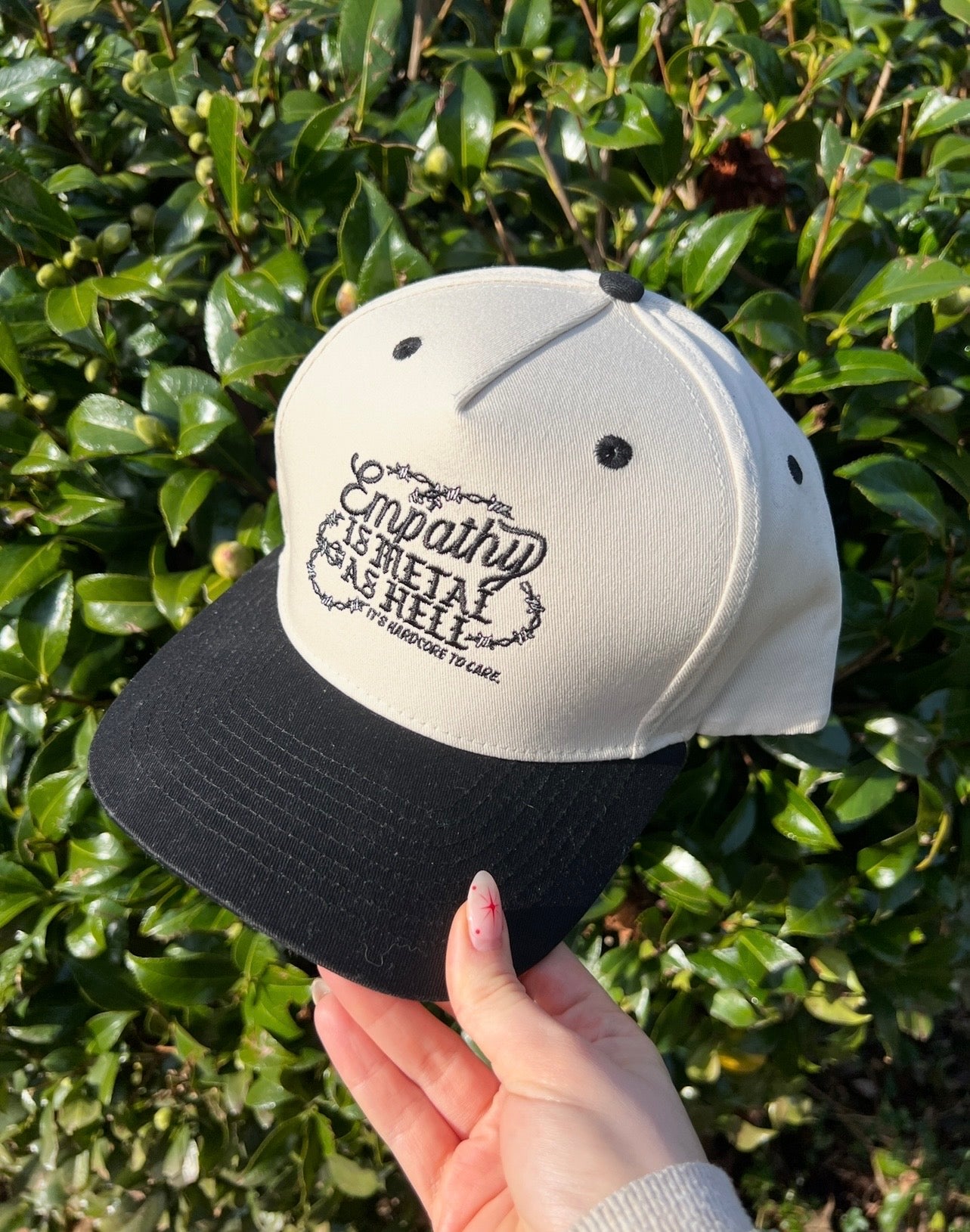 Cap with text that says empathy is metal as hell it's hardcore to care held by a hand against a green leafy background