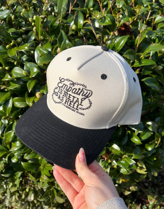Cap with text that says empathy is metal as hell it's hardcore to care held by a hand against a green leafy background