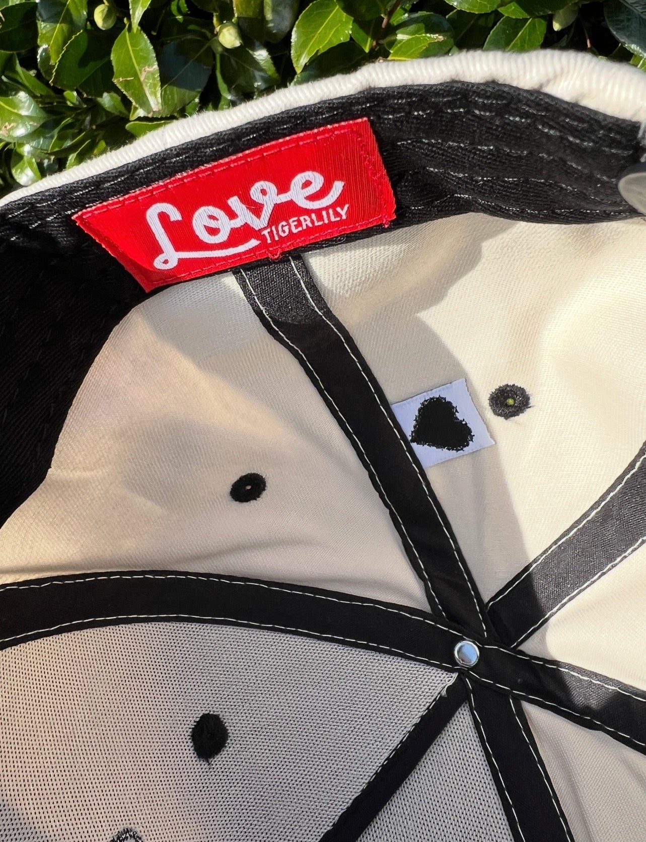 Close-up of a cap with a red 'Love Tigerlily' label and the secret barbed wire heart against a green leafy background