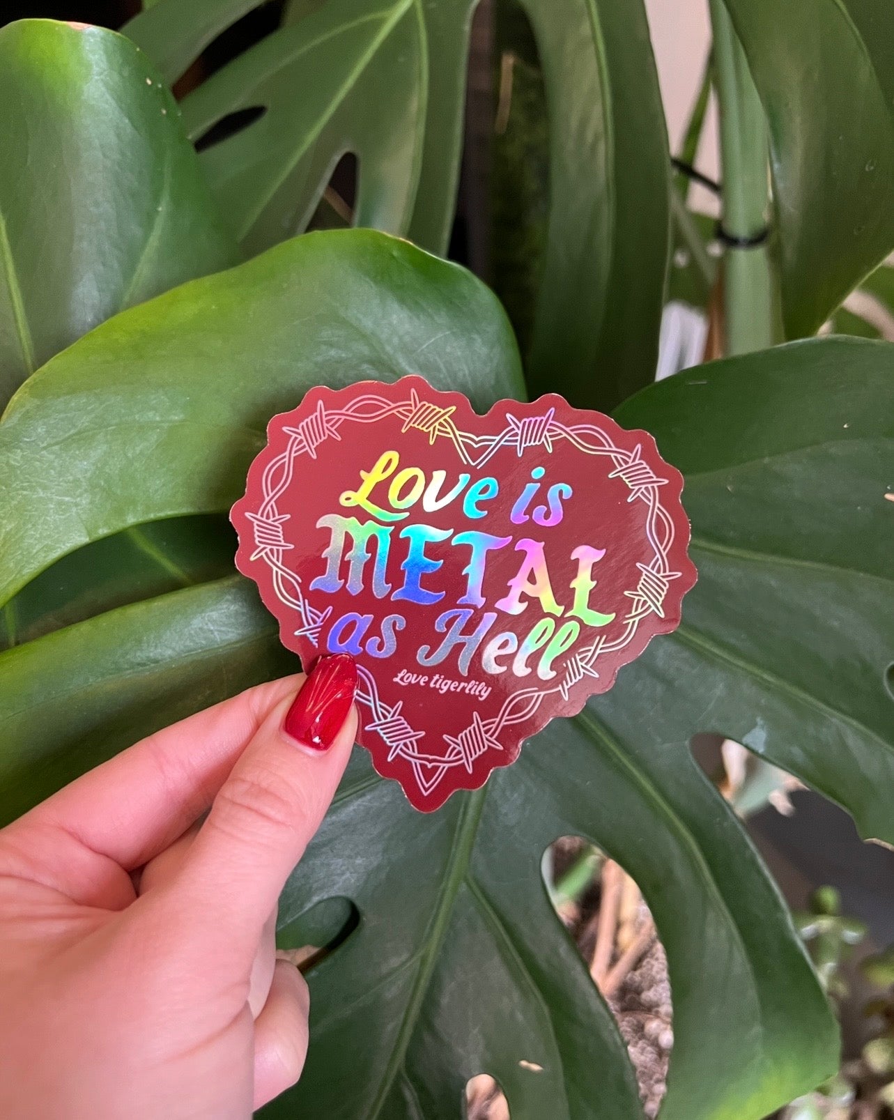 Red Holographic heart stickers with 'Love is metal as hell ' text design with barbed wire held by a monstera plant