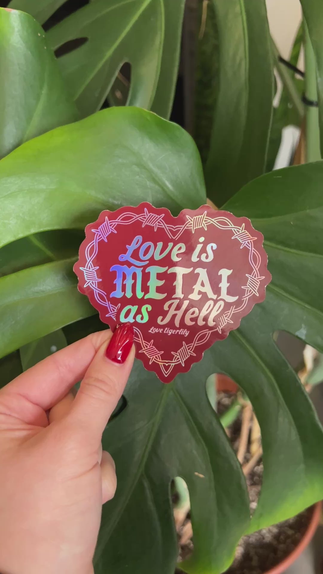 Red Holographic heart stickers with 'Love is metal as hell ' text design with barbed wire held to show off the different rainbow variations