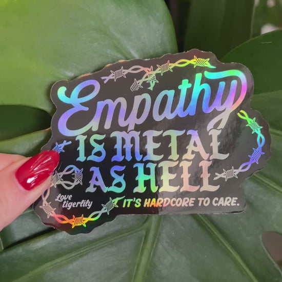 Holographic stickers with 'Empathy is metal as hell & it's hardcore to care' text design with barbed wire held so that it shows the different rainbow variations.