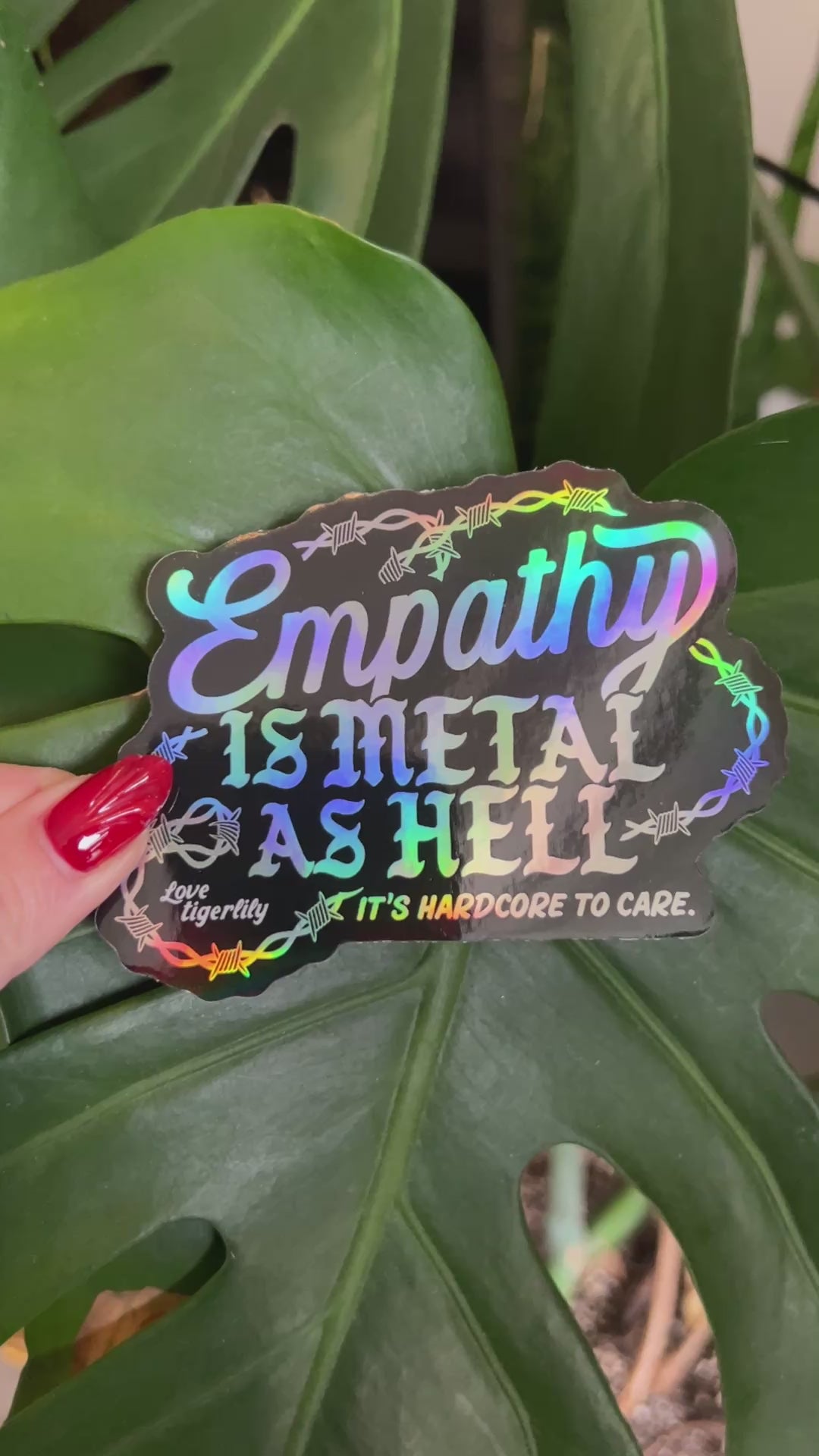 Holographic stickers with 'Empathy is metal as hell & it's hardcore to care' text design with barbed wire held so that it shows the different rainbow variations.