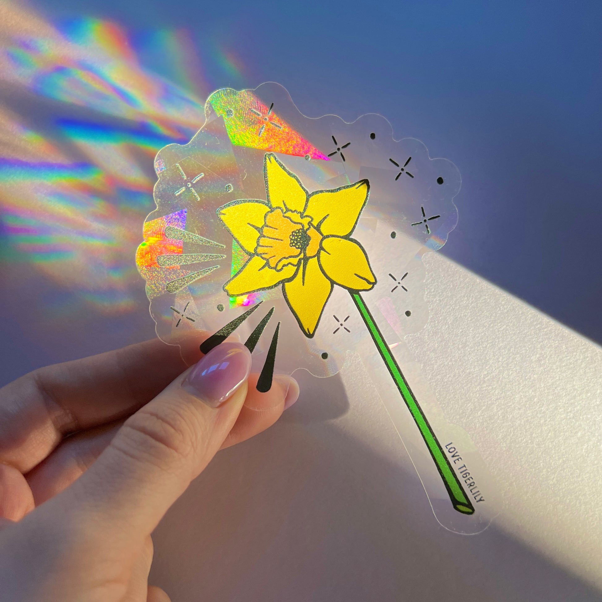 Hand holding the yellow daffodil suncatcher window sticker over a purple background so that it casts rainbows.