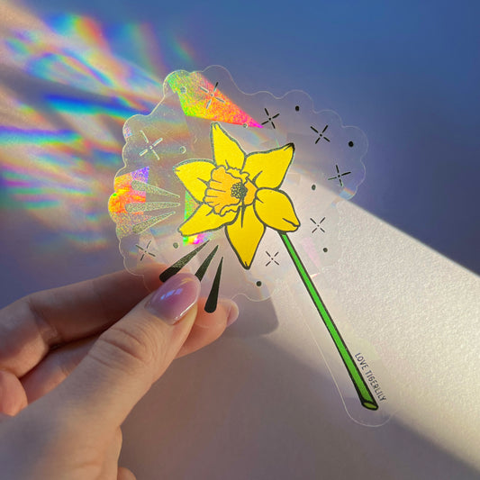 Hand holding the yellow daffodil suncatcher window sticker over a purple background so that it casts rainbows.