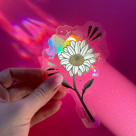 Hand holding the daisy suncatcher window sticker over a pink background so that it casts rainbows.