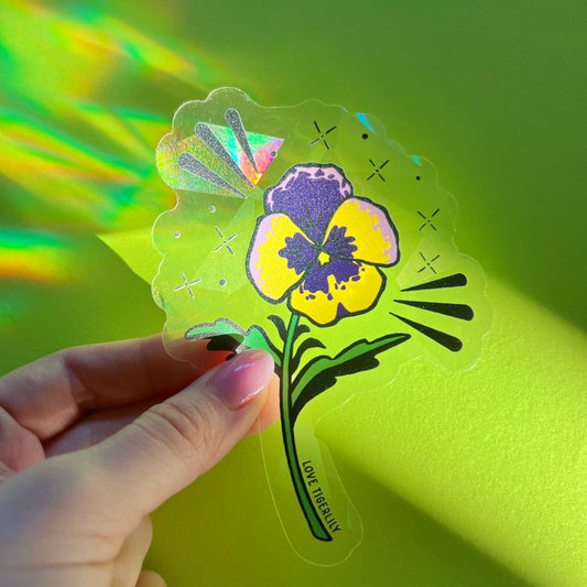 Hand holding the purple and yellow pansy suncatcher window sticker over a lime green background so that it casts rainbows.