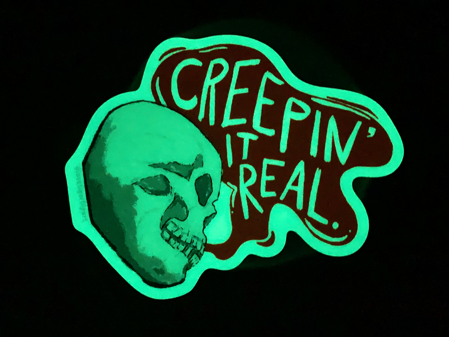 Glow in the Dark Creepin It Real sticker of an illustrated skull saying “Creepin It Real” in the dark glowing green