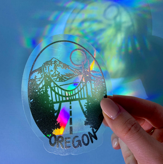 Hand holding the Oregon suncatcher window sticker that is an oval scene with trees, the St. Johns Bridge, and a snowy mountain over a blue background so that it casts rainbows.