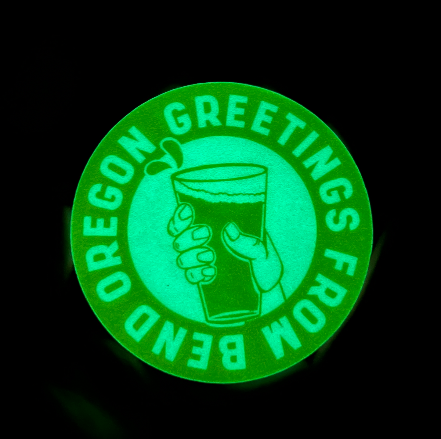 Circular yellow glow in the dark sticker that has an illustrated hand holding a beer with a band of words surrounding the illustration saying “Greetings from Bend Oregon” in the dark and glowing green