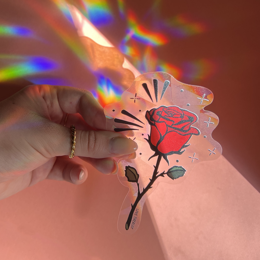 Hand holding the red rose suncatcher window sticker over a pink background so that it casts rainbows.